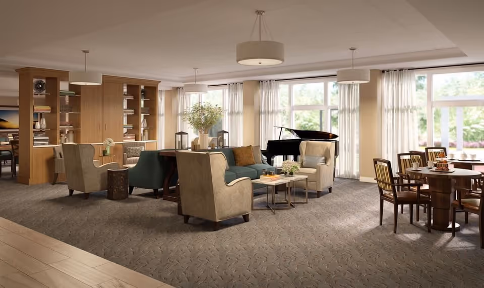 A spacious and well-lit senior living common area with comfortable armchairs and sofas arranged around small tables. Large windows with sheer curtains allow natural light to fill the room. A grand piano is positioned near the windows, and a dining area with wooden tables and chairs is visible on the right. Wooden bookshelves and decorative items are seen in the background.
