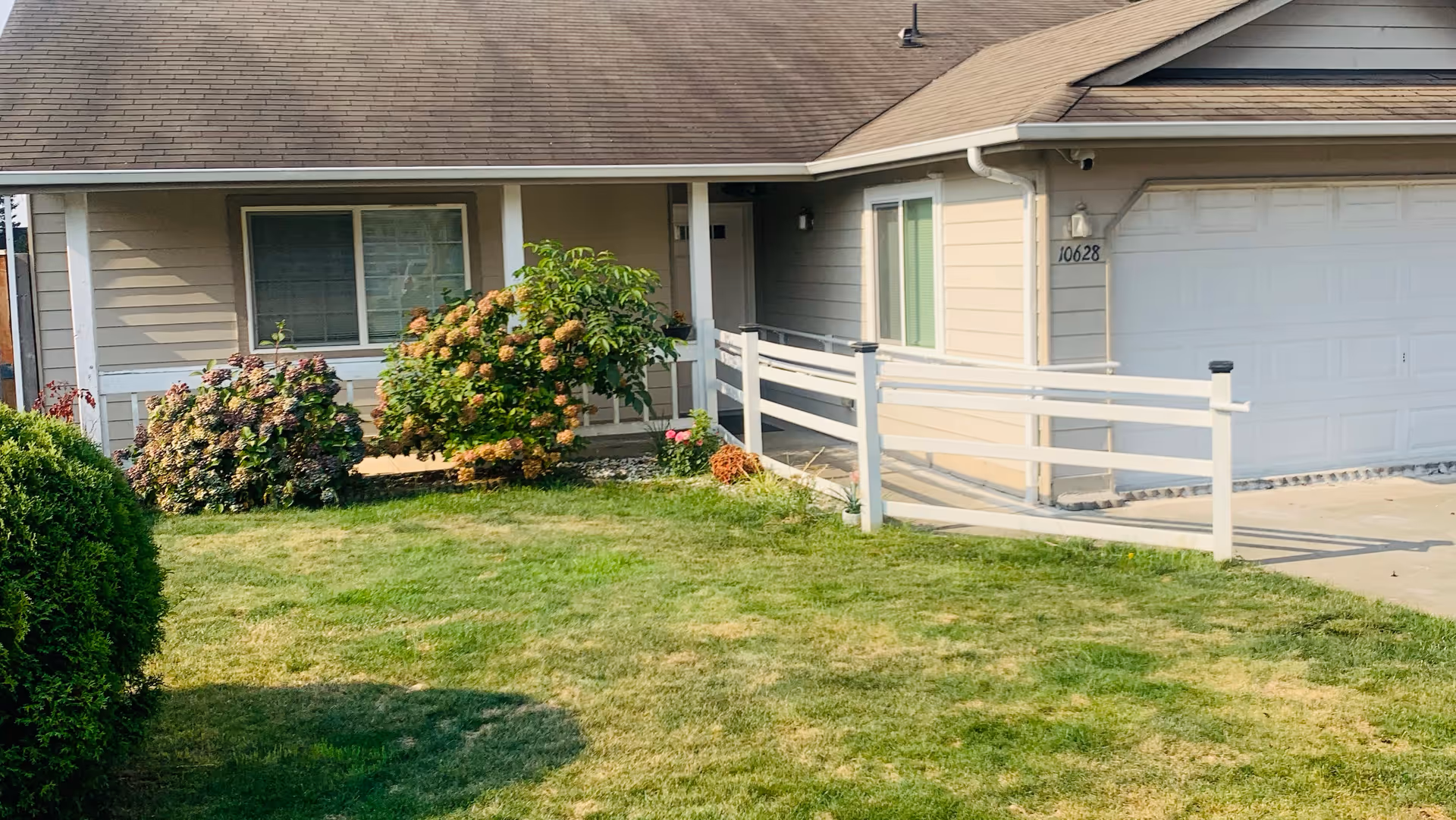 Front exterior view of a single-story residential building with beige siding, a brown shingled roof, a white garage door, and a white fence along the driveway. There are bushes and a green lawn in front of the house.