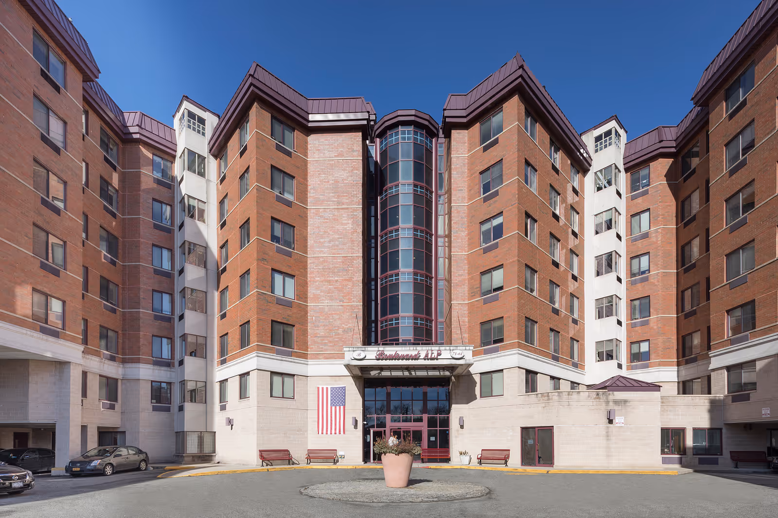 Front exterior view of Boulevard Alp Assisted Living facility, a multi-story brick building with large windows, a central glass entrance, an American flag displayed on the left side, benches near the entrance, and a large flower pot in the circular driveway.
