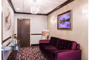 A small waiting or sitting area with a dark purple velvet couch, a glass console table with decorative items, a patterned carpet, and a framed cityscape picture on the wall. The room has beige walls, dark wood trim, and two wall-mounted light fixtures.