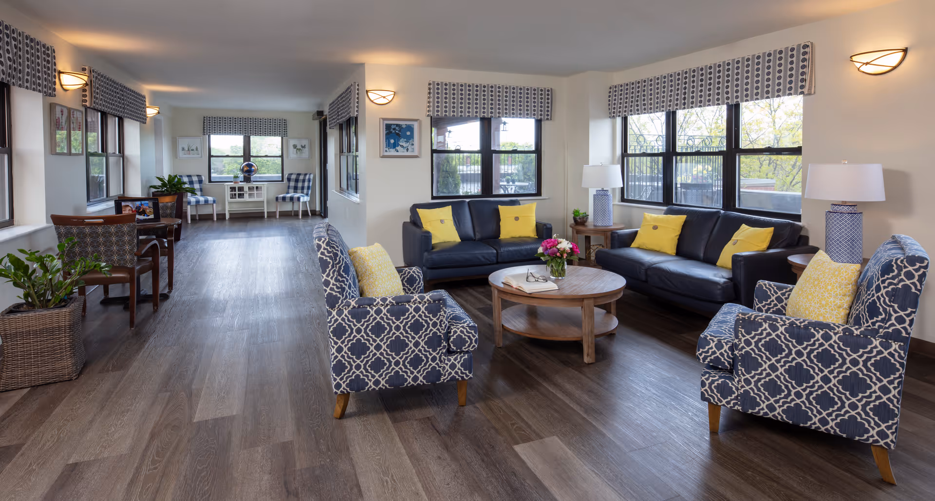 A spacious and well-lit living room area in an assisted living facility featuring two black leather sofas with yellow pillows, two patterned armchairs with yellow cushions, a round wooden coffee table with flowers and a book, large windows with patterned valances, wooden flooring, and additional seating and plants along the walls.