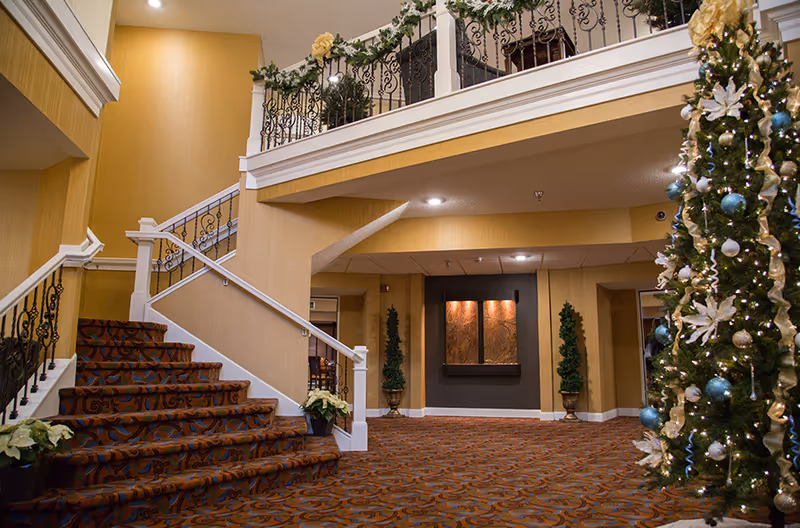 Interior view of a senior living facility lobby with a carpeted staircase on the left, decorated with plants and a railing with ornate ironwork. To the right, a tall Christmas tree adorned with white and blue ornaments and lights. The walls are painted in warm tones, and there is a balcony above with similar railing and greenery decorations.