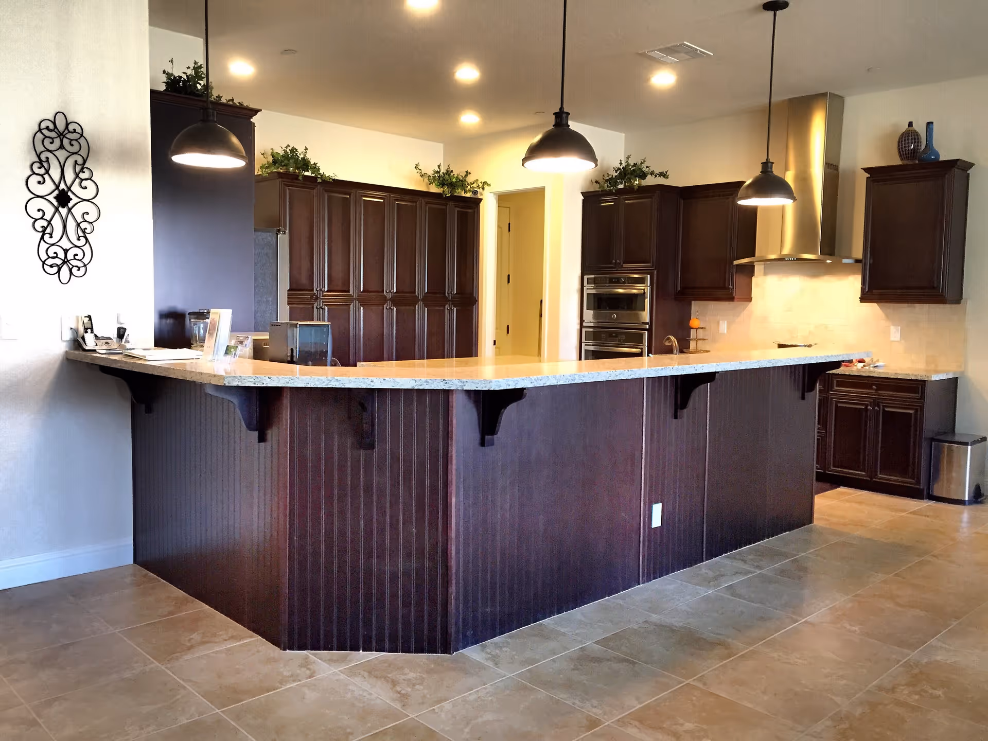 Modern kitchen with dark wood cabinets, a large granite countertop island with pendant lights hanging above, stainless steel appliances including an oven and range hood, and decorative plants on top of the cabinets.