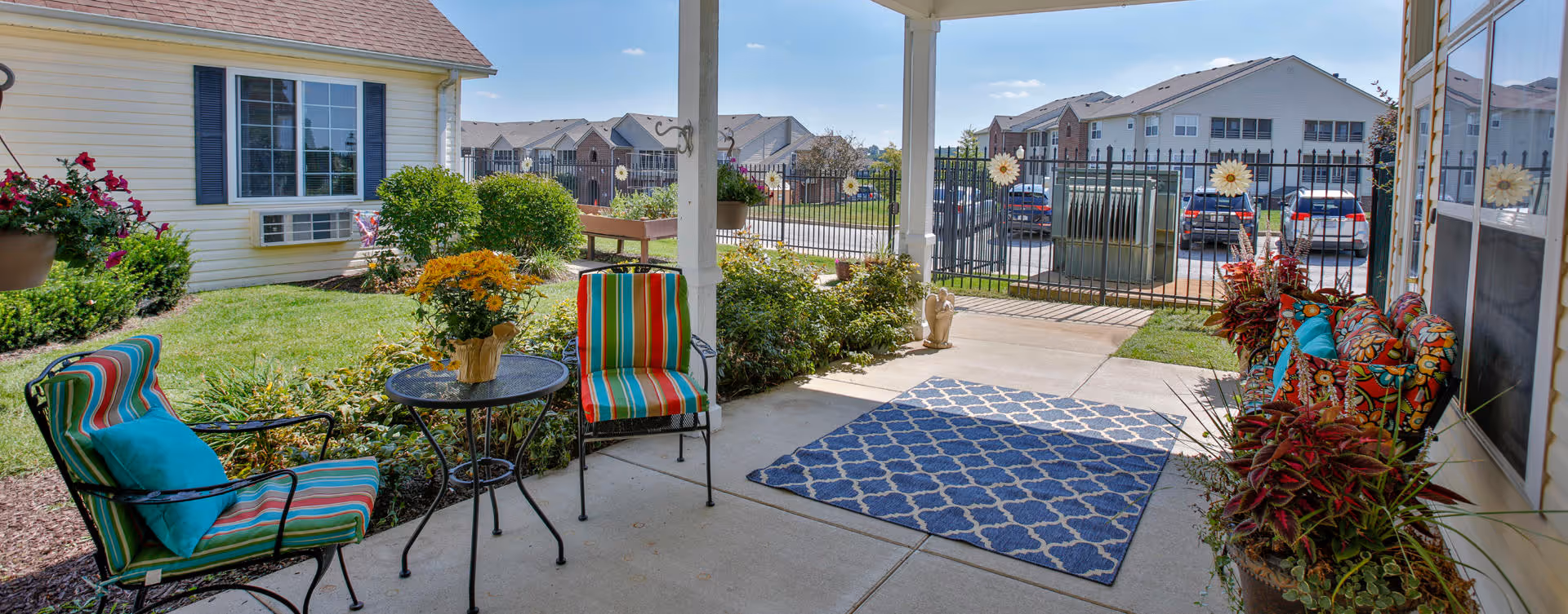 Covered outdoor patio area with colorful striped chairs, a small round table with a flower pot, a blue patterned rug, and various potted plants. In the background, there is a gated parking lot and buildings under a clear blue sky.