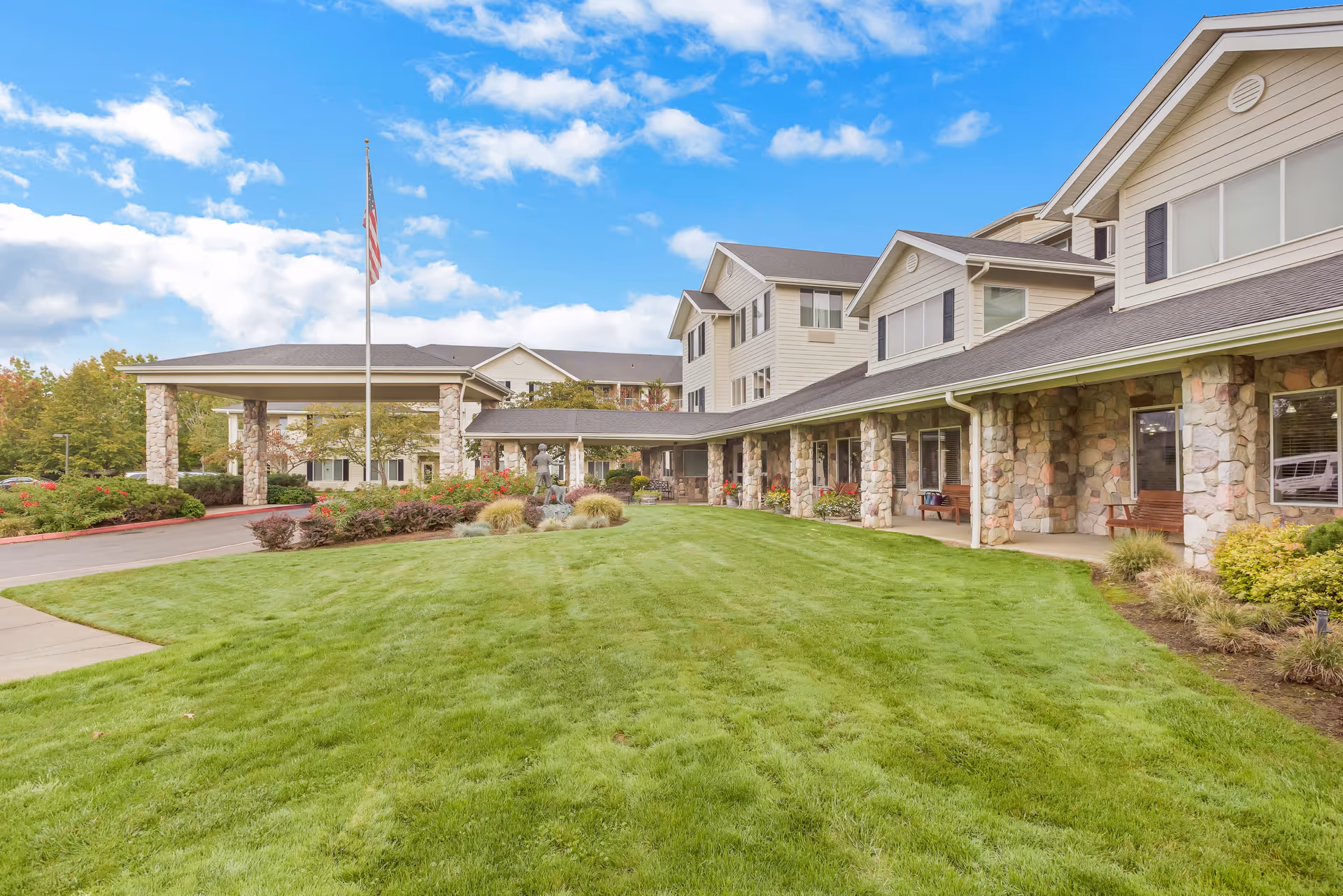 Exterior view of a senior living facility building with stone pillars, a covered entrance, a well-maintained green lawn, and an American flag on a flagpole under a partly cloudy blue sky.