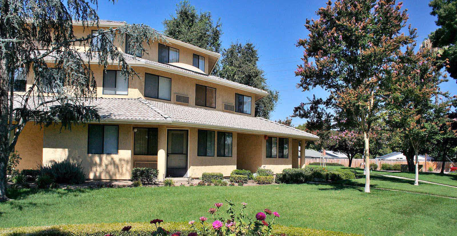 Exterior view of a multi-story senior living community building with beige walls and a tiled roof, surrounded by green grass, bushes, flowering plants, and trees under a clear blue sky.