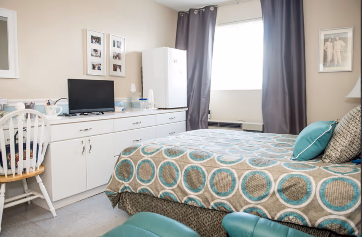 A cozy bedroom with a bed covered in a beige, blue, and brown circular patterned bedspread, two pillows, a white dresser with a small TV, a mini fridge, and a white wooden chair. There are framed photos on the wall and a window with gray curtains letting in natural light.