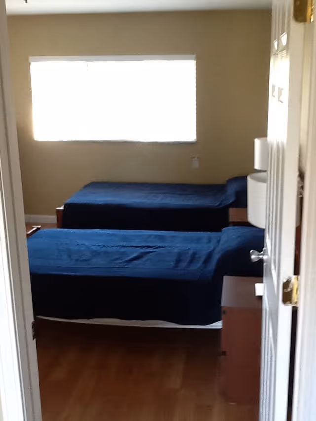 View through a doorway into a bedroom with two single beds covered in dark blue bedspreads. There is a window on the far wall letting in bright light, and a bedside table with a lamp between the beds. The floor is wooden.