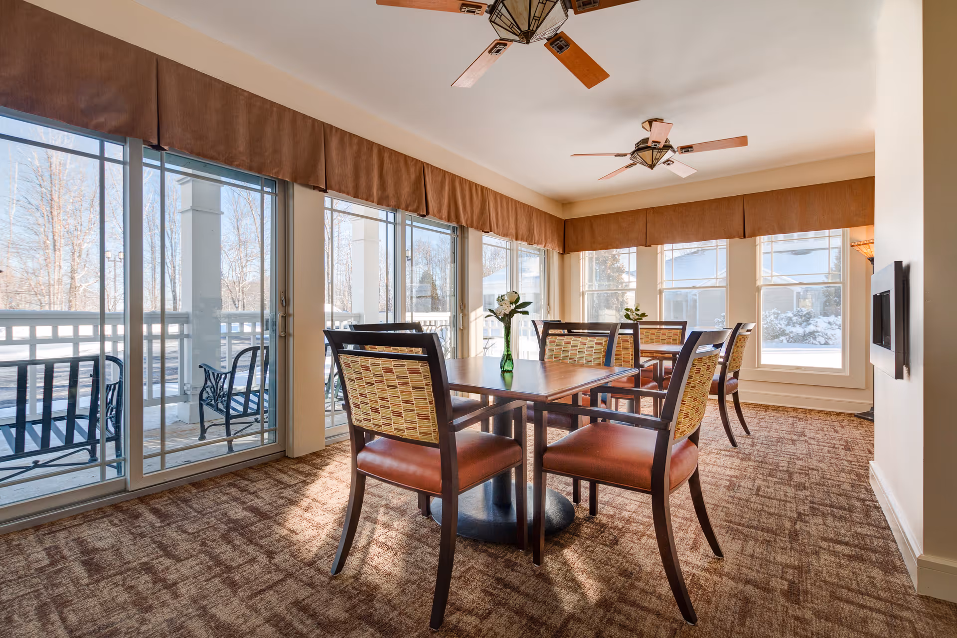A bright dining room with large windows and sliding glass doors showing a snowy outdoor scene. The room has carpeted floors, two ceiling fans with wooden blades, and two square wooden tables each surrounded by four chairs with patterned backs and brown leather seats. A small green vase with white flowers is placed on each table.