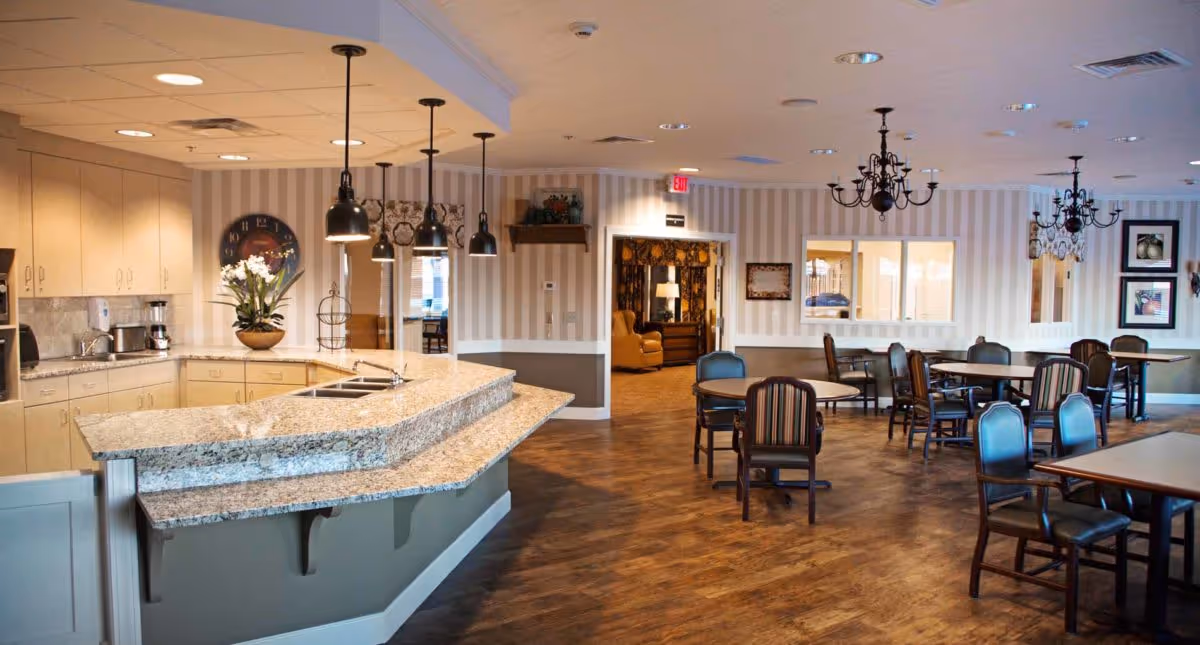 A spacious dining area with multiple tables and chairs arranged on a wooden floor. To the left, there is a kitchen counter with a granite countertop, a sink, and hanging pendant lights above. The walls have striped wallpaper, and there are chandeliers hanging from the ceiling. In the background, a doorway leads to another room with comfortable seating and warm lighting.