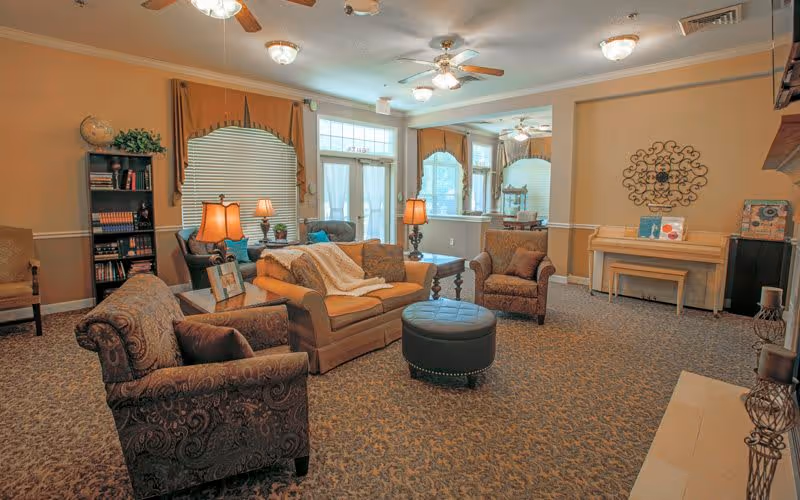 A cozy living room with patterned carpet, a brown sofa with a white throw blanket, two armchairs, a round black ottoman, and several table lamps. There is a bookshelf with books and a globe, a piano with decorative items, and large windows with blinds and curtains letting in natural light.
