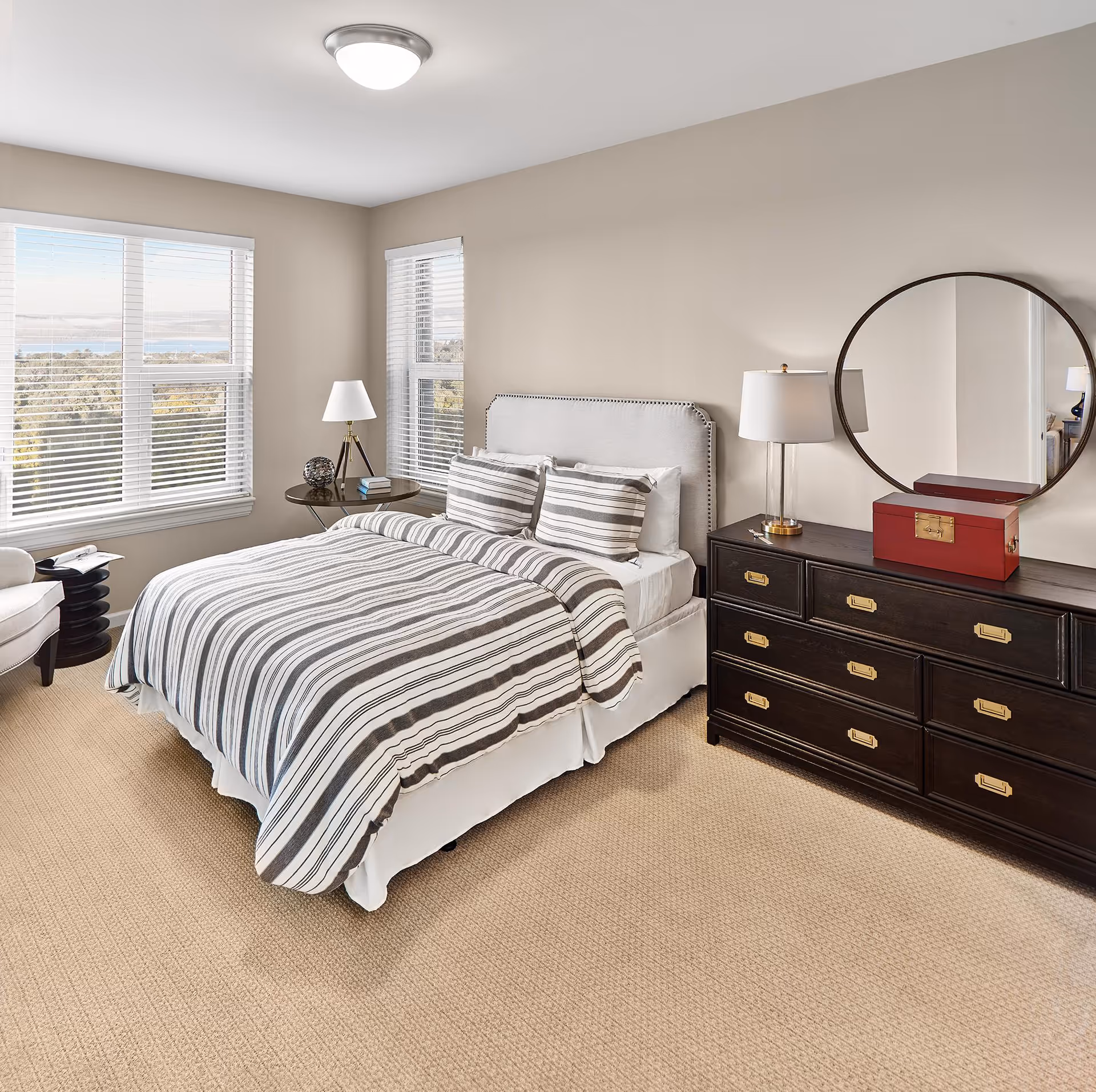 A bright bedroom with a large bed covered in a striped gray and white comforter and matching pillows. The room has beige walls and carpet, two windows with white blinds letting in natural light, a round side table with a lamp and decorative items, a dark wooden dresser with brass handles, a round mirror above the dresser, and a red box on top of the dresser.