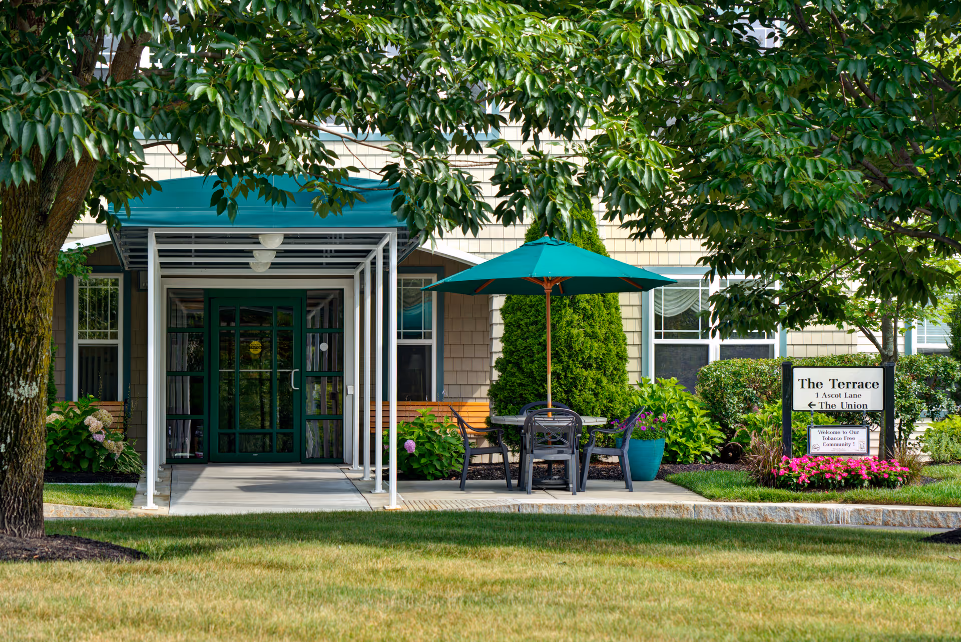 Entrance to a senior living facility named The Terrace At Glen Eddy, featuring a covered walkway, green double doors, outdoor seating with a table and umbrella, surrounded by trees, shrubs, and flowers.