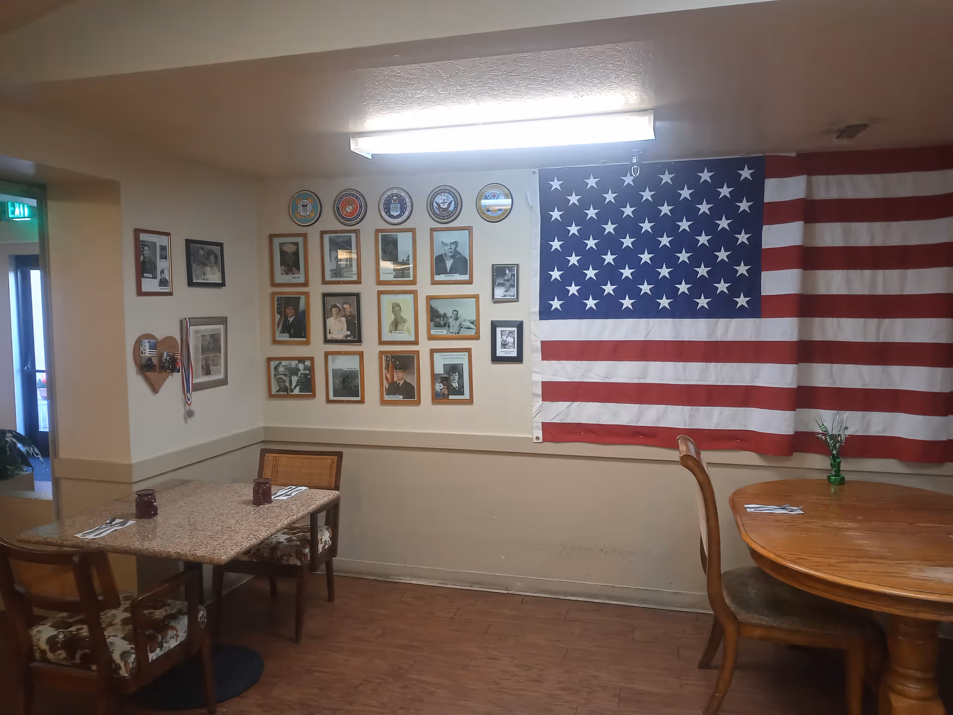 Interior dining area with two tables and chairs. One table is rectangular with a granite-like surface and two chairs with floral upholstery. The other table is round wooden with a single chair. On the wall, there is a large American flag and a collection of framed photographs and military emblems. A fluorescent light fixture is on the ceiling.