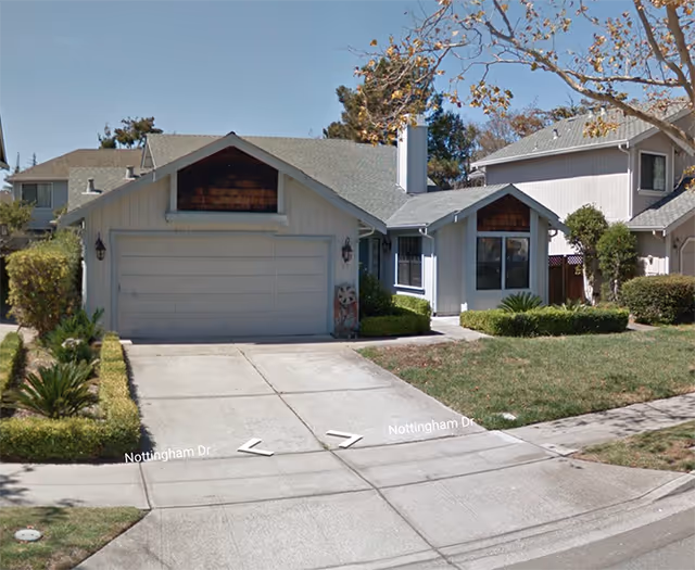 Exterior view of a single-story residential house with a two-car garage, a driveway, and a small front yard with trimmed bushes and a tree. The house has light-colored siding and a shingled roof.