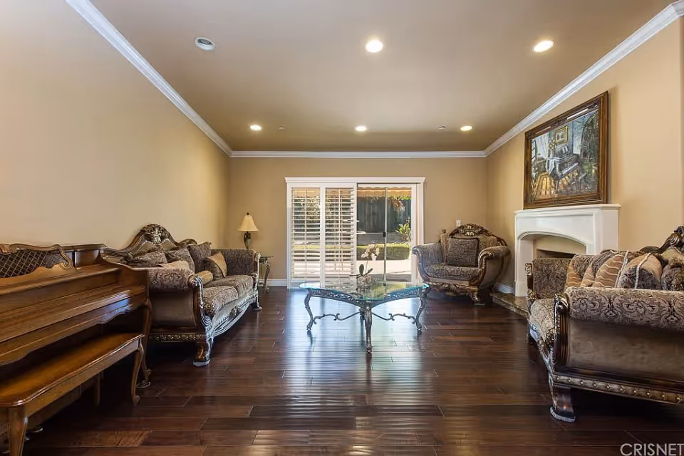 A spacious living room with dark wooden flooring, beige walls, and recessed ceiling lights. The room features ornate upholstered sofas and armchairs arranged around a glass coffee table with a decorative metal base. A wooden piano is positioned on the left side, and a white fireplace with a framed painting above it is on the right. Large sliding glass doors with white plantation shutters lead to an outdoor area.