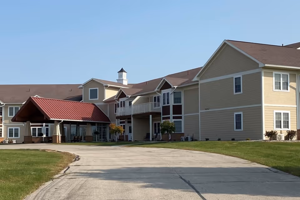 Exterior view of a senior living facility named Garden View Place with beige siding, white trim, and a red-roofed entrance canopy. The building has multiple windows and a driveway leading up to the entrance, surrounded by green grass under a clear blue sky.