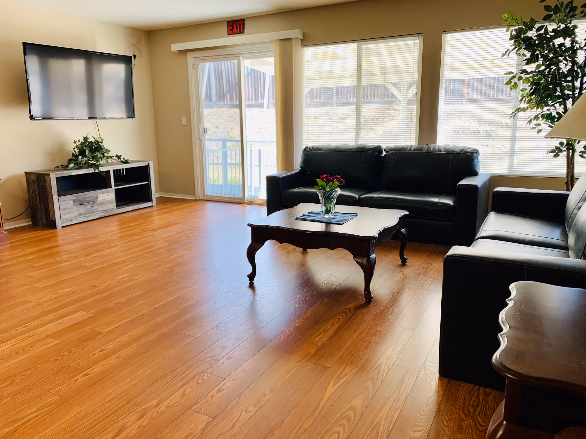 A bright living room with wooden flooring, two black leather sofas, a wooden coffee table with a vase of flowers, a flat-screen TV mounted on the wall above a wooden TV stand, large windows with blinds, a sliding glass door leading outside, and a potted plant in the corner.