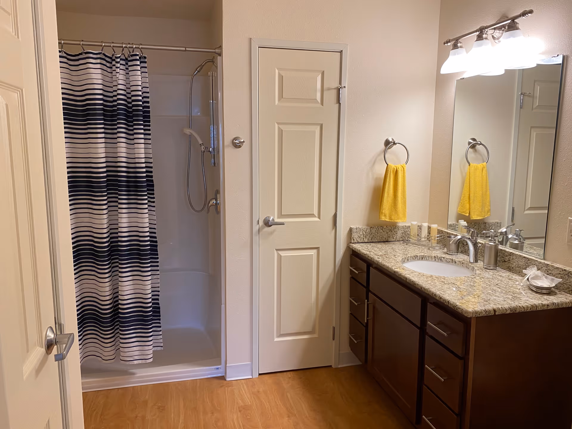 A clean bathroom with a shower area featuring a black and white striped shower curtain, a closed white door, and a vanity with a granite countertop. The vanity has a sink, a silver faucet, soap dispenser, and a yellow hand towel hanging on a ring. Above the vanity is a large mirror with three light fixtures.