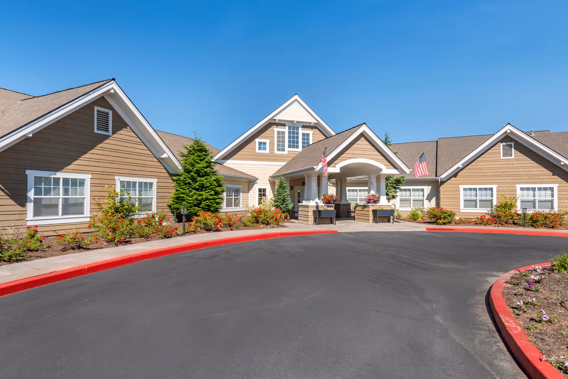 Front exterior view of a senior living facility building with beige siding, white trim, and a covered entrance. There are American flags on either side of the entrance, landscaped flower beds with red and pink flowers, and a clear blue sky above.