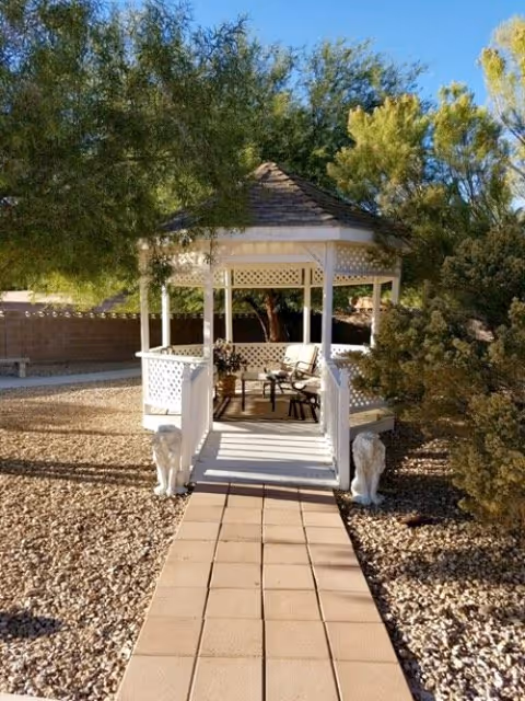 A white wooden gazebo with a shingled roof situated in a garden area with gravel ground cover and surrounded by trees and bushes. A paved walkway leads up to the gazebo entrance, which is flanked by two white lion statues. Inside the gazebo, there are chairs and a small table.