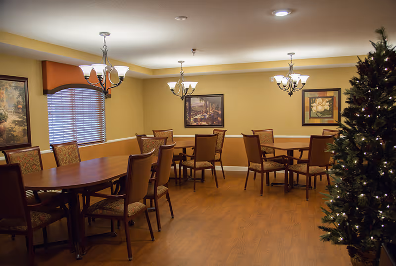 A dining room with wooden floors, several wooden tables and chairs with patterned upholstery, three chandeliers hanging from the ceiling, framed artwork on the walls, a window with blinds and an orange valance, and a decorated Christmas tree on the right side.