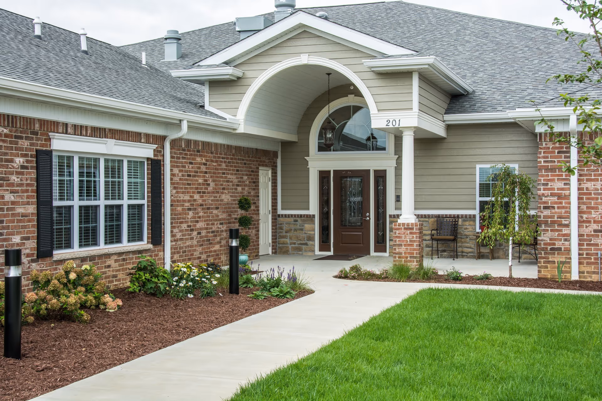 Front entrance of a senior living facility with a covered porch, brick and siding exterior, a brown door with glass panels, windows with black shutters, landscaped garden beds, and a concrete walkway leading to the door.