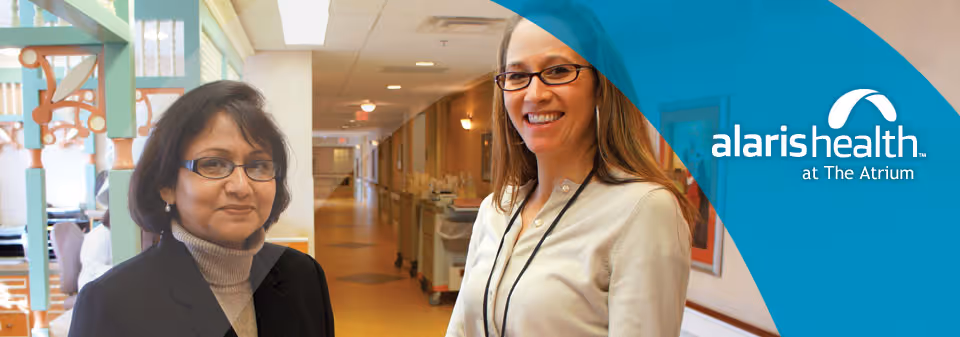 Two women standing in a well-lit hallway of a healthcare facility, one wearing glasses and a black blazer, the other smiling and wearing glasses and a light-colored top with a lanyard. The hallway has medical equipment and doors along the walls.