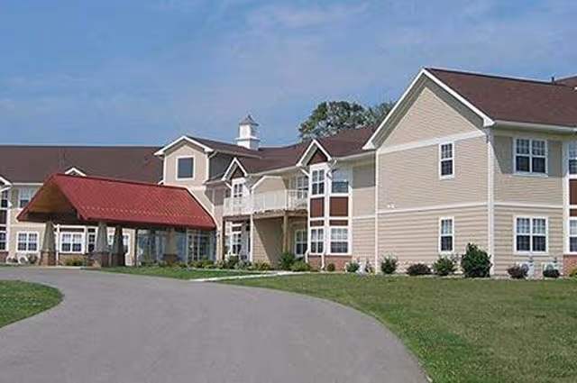 Exterior view of a two-story senior living facility building with beige siding and a red roof over the entrance. The building is surrounded by a paved driveway and green lawn under a clear blue sky.