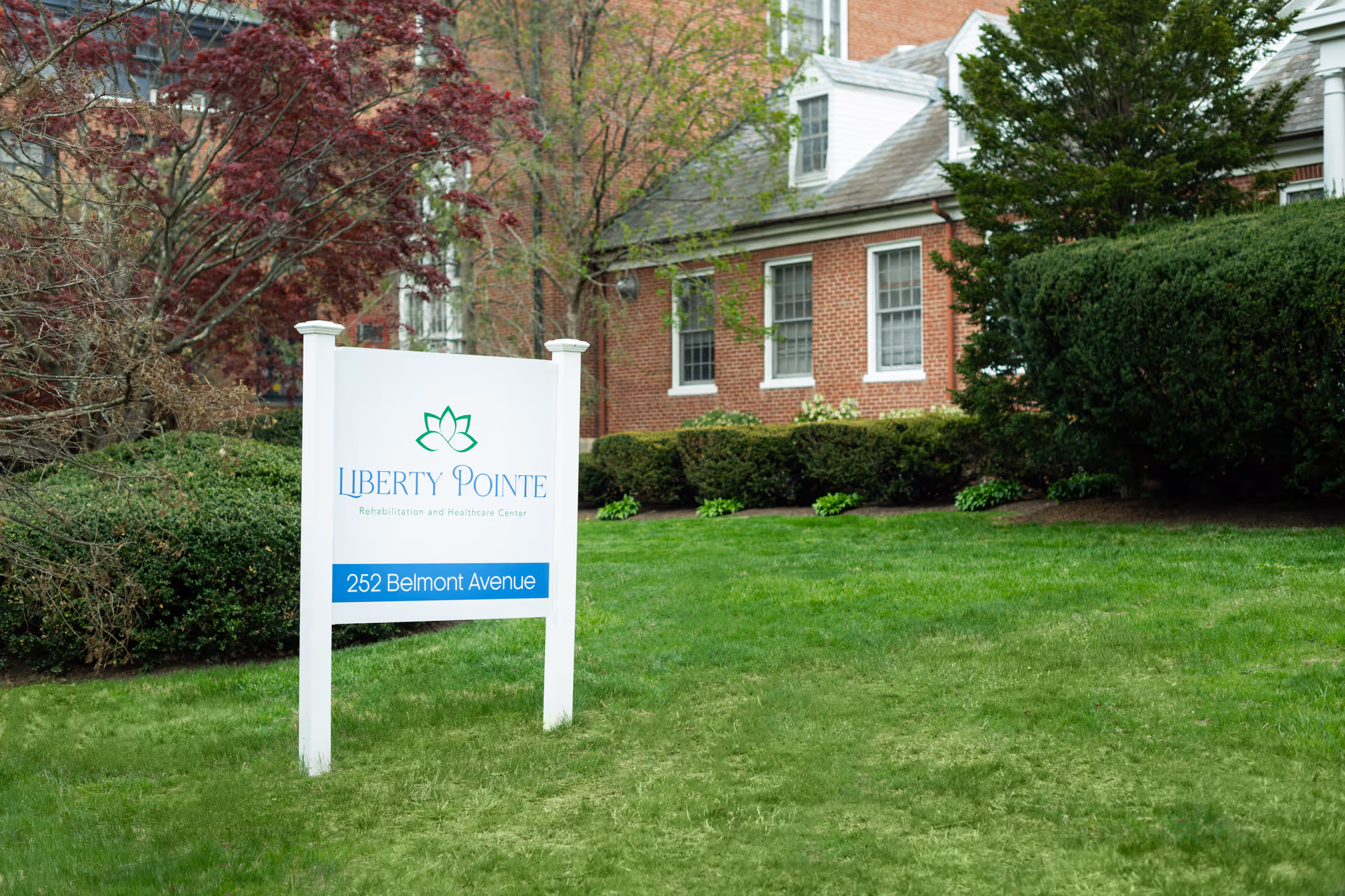 A white sign with green and blue text stands on a well-maintained grassy lawn in front of a brick building. The sign reads 'Liberty Pointe Rehabilitation and Healthcare Center' with the address '252 Belmont Avenue'. Surrounding the sign are bushes and trees with some red and green foliage.
