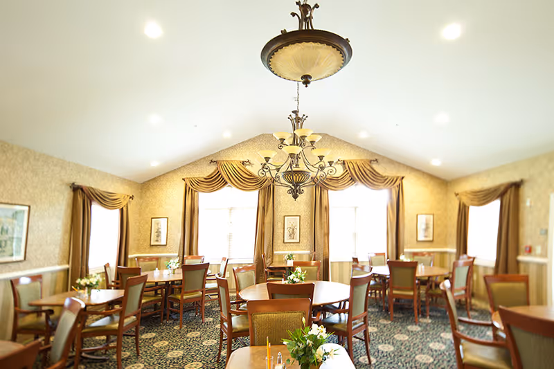 A bright dining room with multiple round and rectangular wooden tables surrounded by chairs with green upholstery. The room features large windows with elegant gold curtains and valances, patterned carpet, and a decorative chandelier hanging from the ceiling. Small floral centerpieces are placed on the tables.