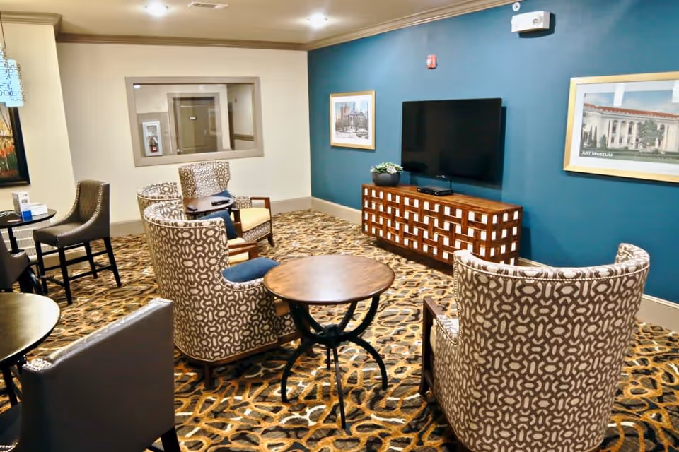A cozy lounge area with patterned armchairs arranged around small round wooden tables. A flat-screen TV is mounted on a teal accent wall above a wooden cabinet. The room has a patterned carpet and framed artwork on the walls.