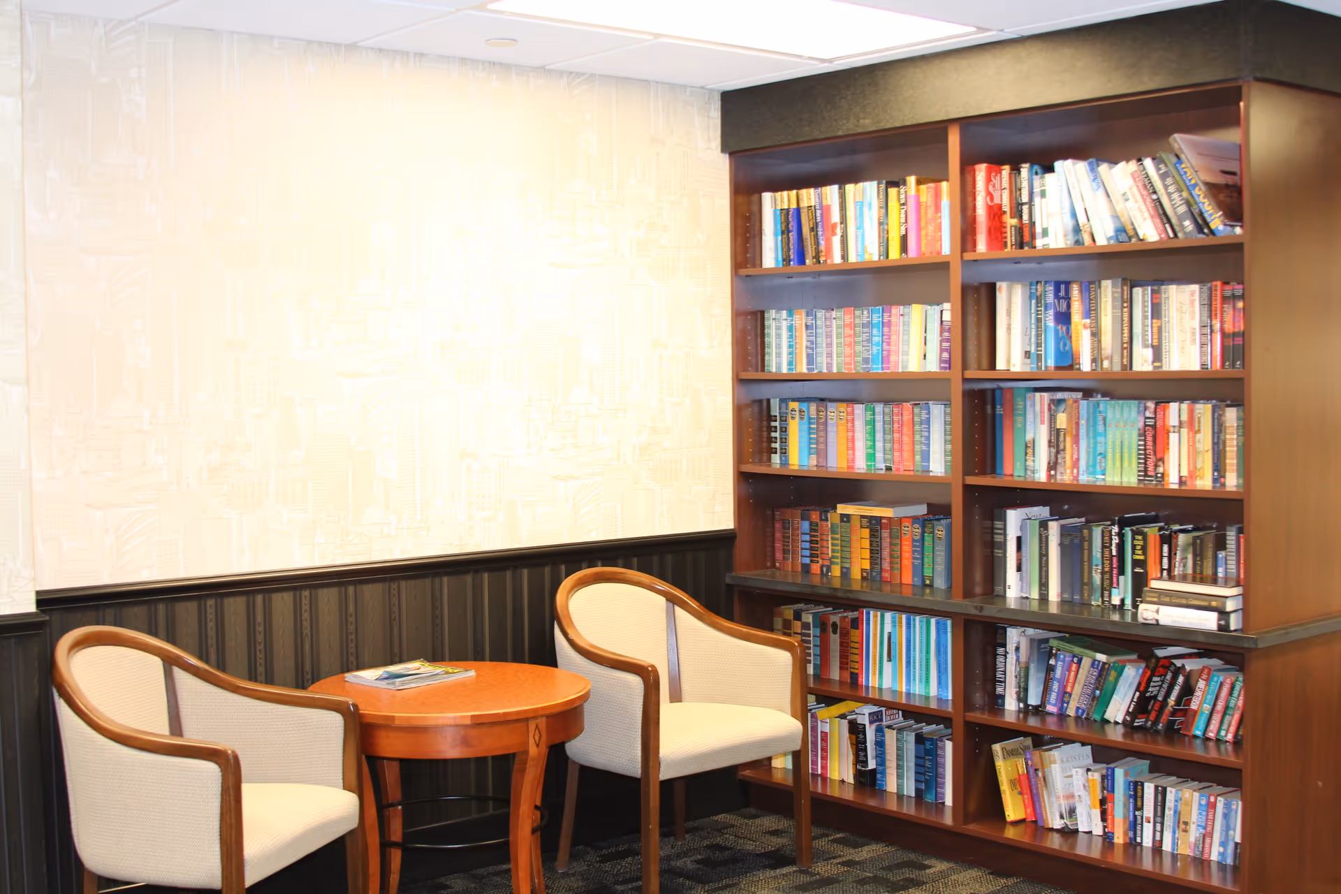 A cozy reading nook with two beige upholstered chairs and a round wooden table between them. Behind the chairs is a large wooden bookshelf filled with various books. The wall has a light-colored wallpaper with a subtle cityscape pattern and dark wainscoting below.