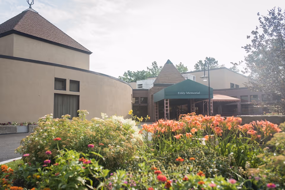 Exterior view of the Terrace At Eddy Memorial building with a garden full of colorful flowers in the foreground and a green awning above the entrance that reads 'Eddy Memorial'.