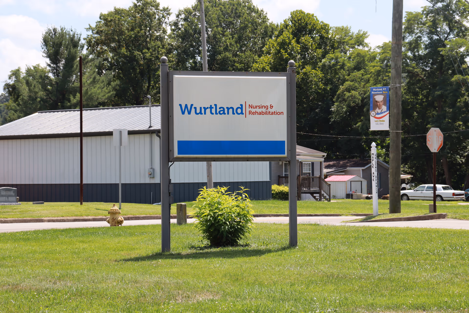 Outdoor view of a sign for Wurtland Nursing & Rehabilitation on a grassy area with trees and buildings in the background under a partly cloudy sky.