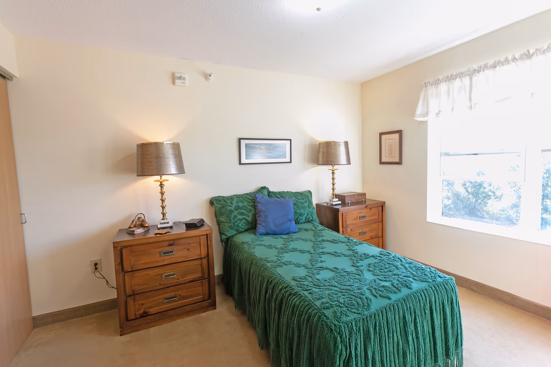 A small bedroom with a single bed covered in a green textured bedspread and a blue pillow. There are two wooden nightstands on either side of the bed, each with a brass lamp. A framed picture hangs on the wall above the bed, and a window with white curtains lets in natural light on the right side of the room.