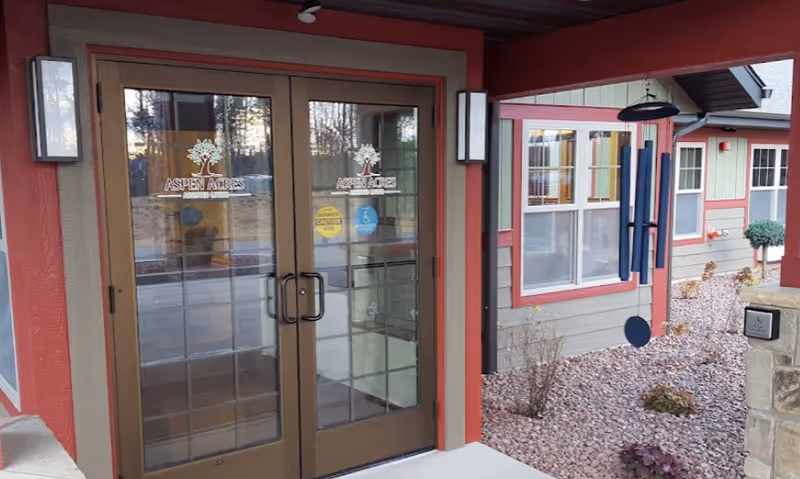 Entrance to Aspen Acres Assisted Living facility with double glass doors featuring the facility's logo. The exterior walls are painted in red and beige tones, with windows and a landscaped area with rocks and small plants. A large blue wind chime hangs near the entrance.