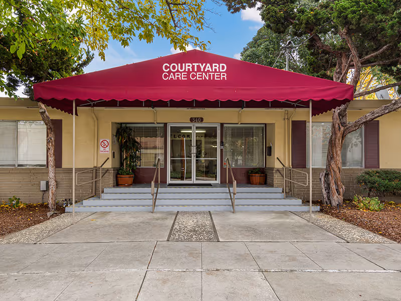 Front entrance of Courtyard Care Center with a red canopy displaying the facility name. The entrance has steps and handrails leading to glass double doors, flanked by windows and potted plants. Trees and landscaping are visible on either side.