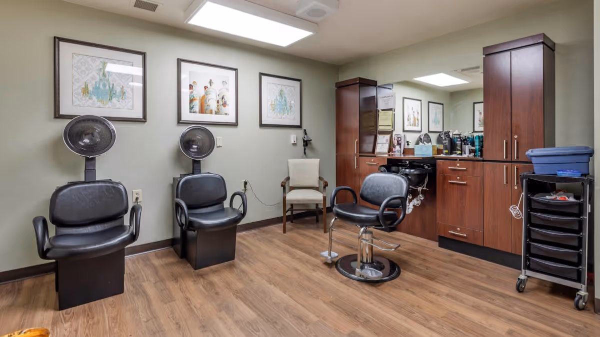 Interior view of a salon area in a senior living facility with three black salon chairs, two with hair dryers attached. The room has wooden flooring, light green walls, framed artwork on the walls, and wooden cabinets with a countertop holding various hair care products. There is also a rolling cart with drawers and a blue container on top.