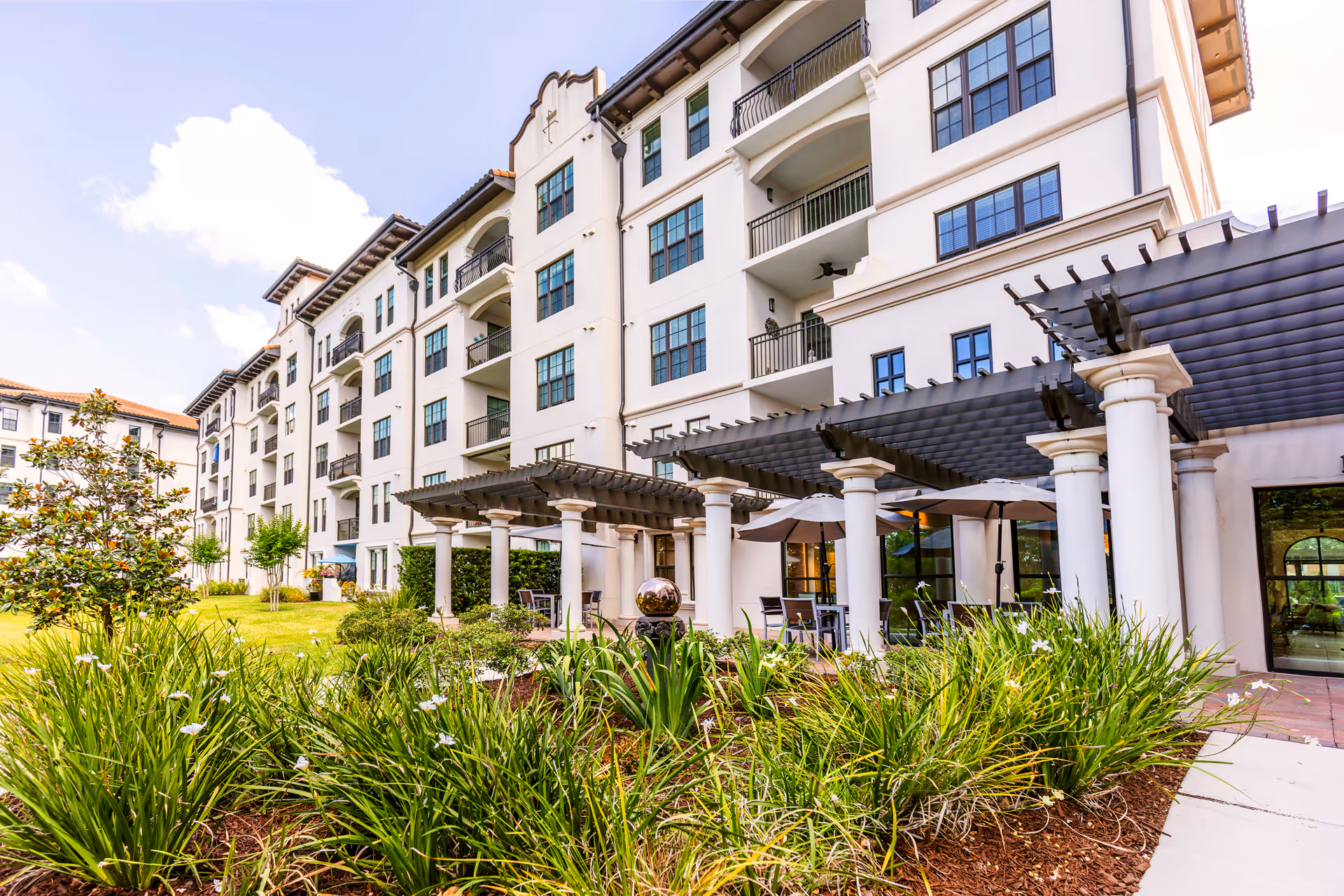 Exterior view of a multi-story senior living facility building with balconies, a pergola-covered patio area with tables and umbrellas, and landscaped greenery in the foreground under a partly cloudy sky.