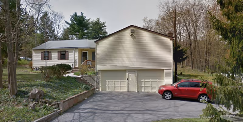 Exterior view of a single-story residential building with beige siding, two garage doors, a small front porch, and a red SUV parked in the driveway. The surrounding area has trees and greenery.