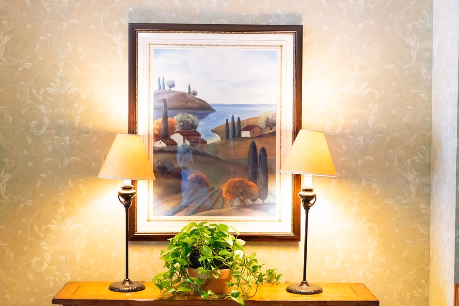 A wooden table with a green potted plant on it, flanked by two lit table lamps with beige lampshades. Above the table hangs a framed landscape painting depicting hills, trees, and a body of water under a cloudy sky. The background wall has a light beige floral patterned wallpaper.