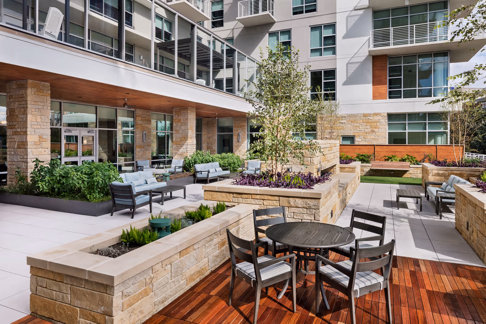 Outdoor patio area at The Village at The Triangle featuring modern seating arrangements with cushioned chairs and tables, surrounded by stone planters with greenery and small trees, adjacent to a multi-story building with large windows and balconies.