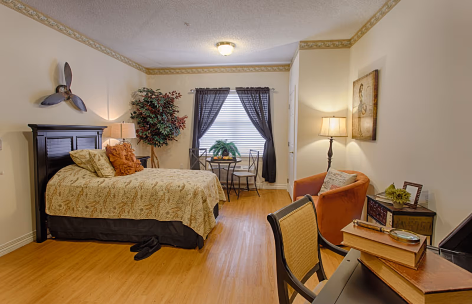 A cozy bedroom in a senior living facility featuring a bed with patterned bedding and decorative pillows, a small round table with two chairs near a window with dark curtains, a potted plant in the corner, an orange armchair with a floor lamp beside it, and a desk with books and a magnifying glass on top.