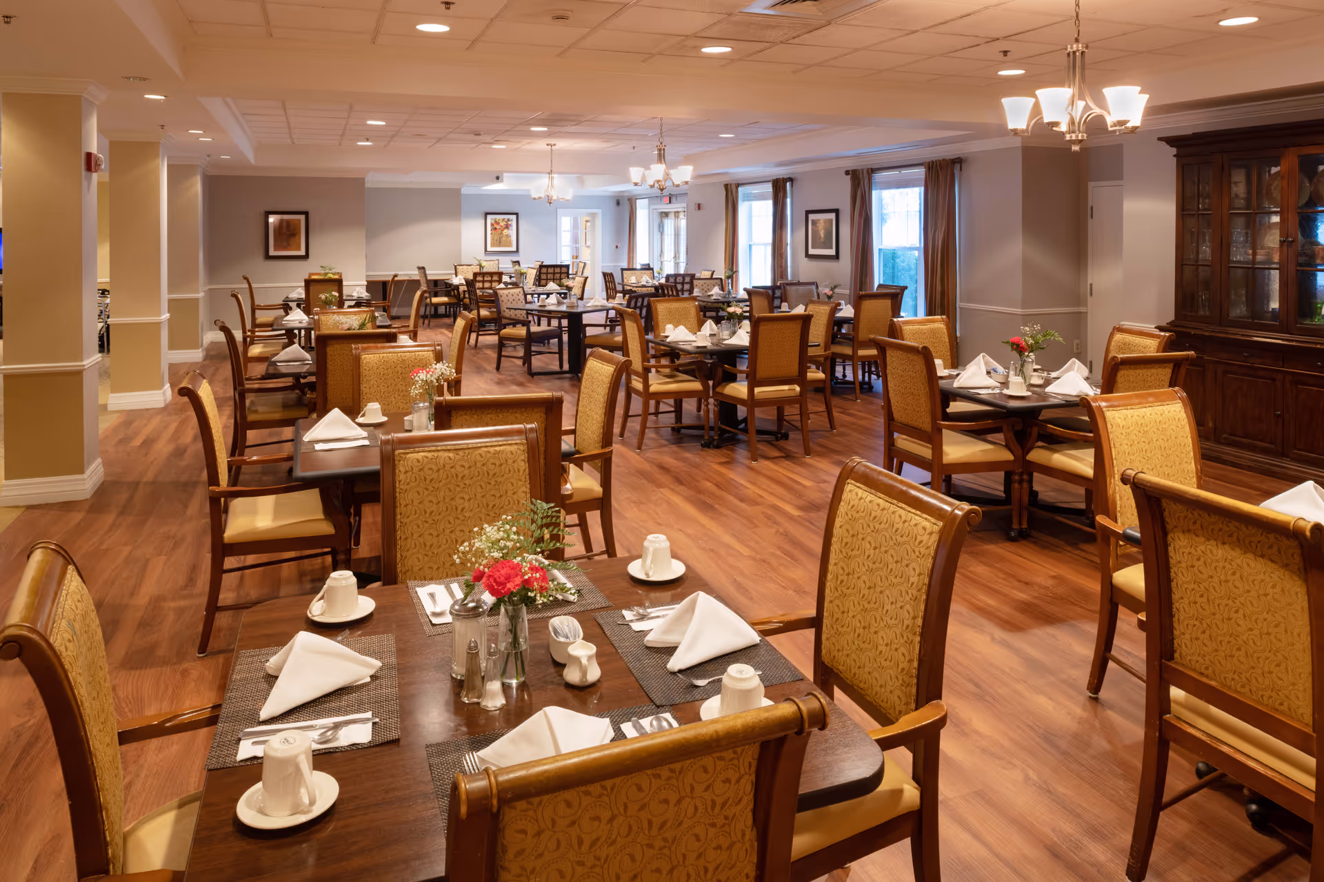 A spacious dining room in a senior living facility with multiple tables set for meals. Each table has neatly folded white napkins, cups, and small flower arrangements. The room features wooden flooring, cushioned chairs with patterned upholstery, soft lighting from chandeliers, and large windows with curtains allowing natural light.