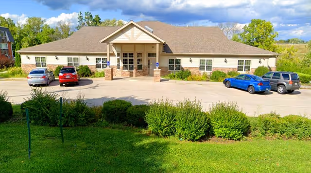 Exterior view of a single-story building with a peaked entrance and a parking lot in front. Four cars are parked in the lot, and there are green bushes and grass in the foreground under a partly cloudy sky.