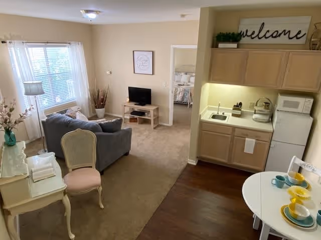 A cozy living space featuring a small kitchenette with a sink, microwave, and refrigerator on the right. A round dining table set with colorful dishes is in the foreground. To the left, there is a sitting area with a gray sofa, a cushioned chair, a floor lamp, and a small TV on a wooden stand. A window with white curtains lets in natural light. In the background, a doorway leads to a bedroom with a bed and pillows visible. The walls are painted beige, and there is a decorative 'welcome' sign above the kitchenette.