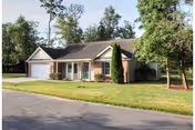 Single-story house with a two-car garage, surrounded by green grass and trees under a clear sky.