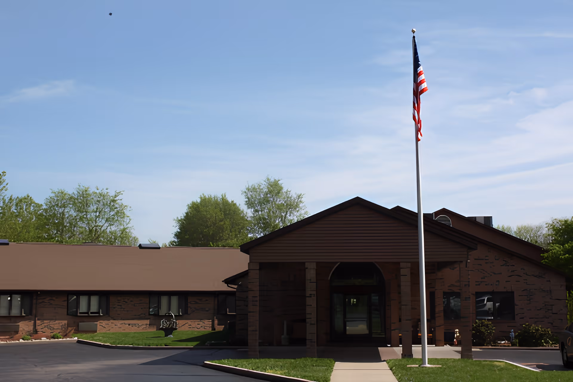 Exterior front view of a single-story senior living facility building with a covered entrance and an American flag on a flagpole in front. The building is brown with several windows and surrounded by green trees and grass under a clear blue sky.