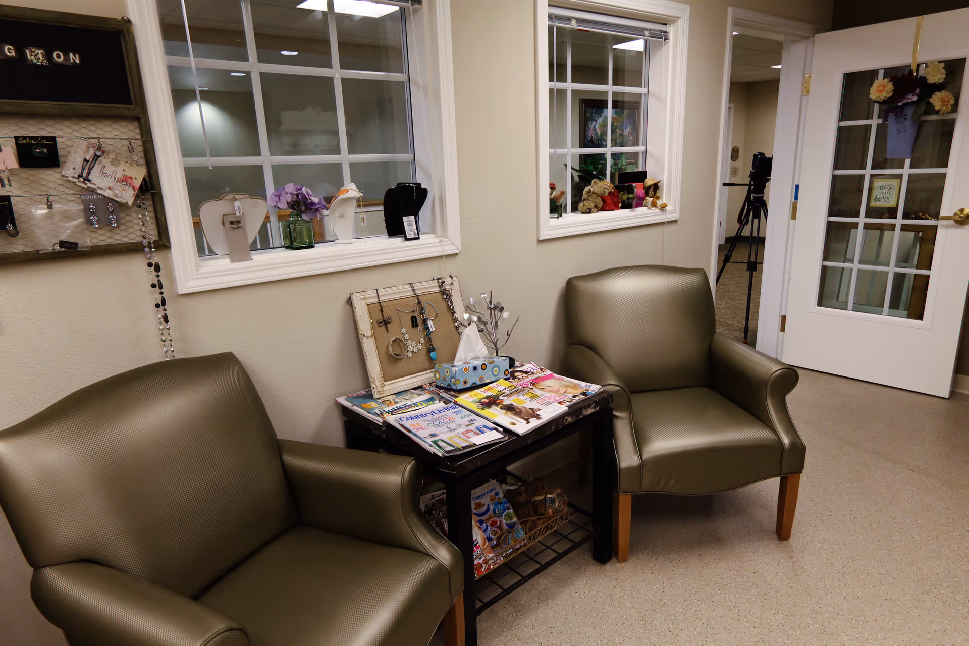 A small waiting area with two green leather armchairs and a black side table between them. The table holds magazines, a tissue box, and a jewelry display. Behind the chairs are two windows with white frames looking into another room, and a white door with glass panes is visible on the right side.